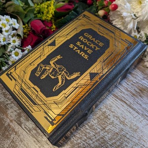 Puede incluir: Un libro de tapa dura negro con detalles dorados. El t&iacute;tulo "GRACE ROCKY SAVE STARS" est&aacute; impreso en oro. La portada presenta un dise&ntilde;o abstracto. El libro est&aacute; rodeado de flores.