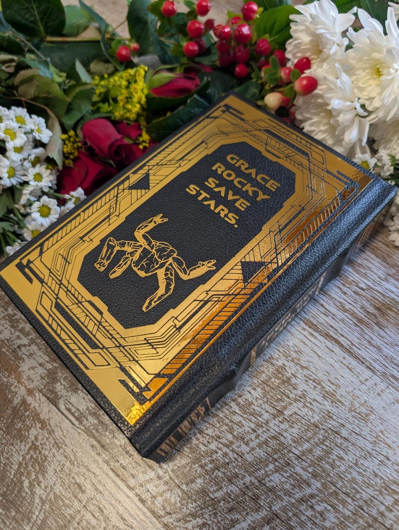 Puede incluir: Un libro de tapa dura negro con detalles dorados. El t&iacute;tulo "GRACE ROCKY SAVE STARS" est&aacute; impreso en oro. La portada presenta un dise&ntilde;o abstracto. El libro est&aacute; rodeado de flores.