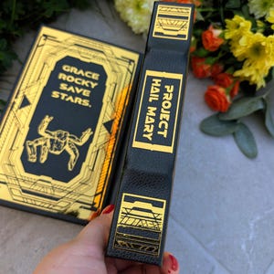 Puede incluir: Un libro negro con detalles dorados y el t&iacute;tulo "Project Hail Mary" en el lomo. La portada presenta el texto "Grace Rocky Save Stars" y una ilustraci&oacute;n estilizada. El libro se sostiene cerca de un ramo de flores.