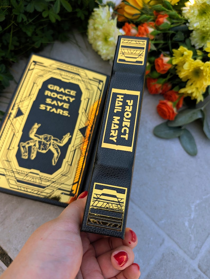 Puede incluir: Un libro negro con detalles dorados y el t&iacute;tulo "Project Hail Mary" en el lomo. La portada presenta el texto "Grace Rocky Save Stars" y una ilustraci&oacute;n estilizada. El libro se sostiene cerca de un ramo de flores.