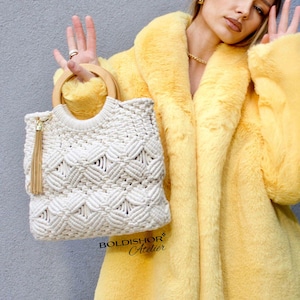 以下が含まれることがあります： 木製のハンドルと金のタッセルが付いた、クリーム色のマクラメハンドバッグ。 バッグは幾何学模様で、長い黄色のフェイクファーコートを着た人の前に持たれています。 テキスト「BOLDISHOR Atelier」が表示されています。
