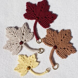Può includere: Quattro ornamenti a forma di foglia in macramè fatti a mano nei toni del bordeaux, beige, marrone e giallo. Ogni foglia presenta uno stelo con una piccola perla di legno e un anello dorato. Le foglie sono disposte su una superficie bianca strutturata.