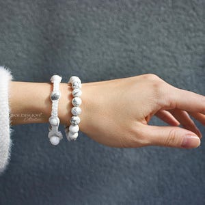 May include: Two white beaded bracelets are worn on a wrist, one with a macrame design and the other with round beads. The bracelets are white with gray accents. The background is a solid gray color.