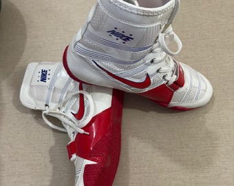 Könnte beinhalten: Weiße und rote High-Top-Sportschuhe mit dem Nike-Logo. Die Schuhe haben einen roten Swoosh und Akzente, weiße Schnürsenkel und einen Riemen. Die Sohlen sind rot und strukturiert. Für sportliche Aktivitäten konzipiert.