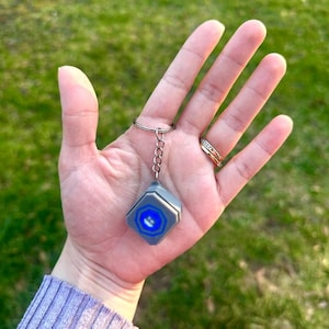 May include: A silver keychain with a small, gray, hexagonal charm. The charm has a blue logo with a white outline.