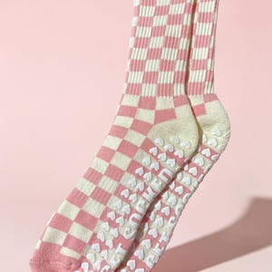 May include: A pair of pink and white chequered socks. The socks feature a pink toe and heel, with a white heart-patterned grip on the sole. The background is a solid pink.