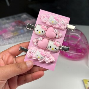 May include: Three silver-toned metal hair clips attached to a pink display card. Each clip features a white Hello Kitty character face, pink heart accents, and decorative pink swirls resembling frosting, embellished with small clear rhinestones.