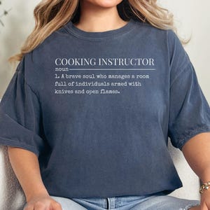 May include: A navy blue t-shirt with the text "COOKING INSTRUCTOR" and a definition. The definition reads: "1. A brave soul who manages a room full of individuals armed with knives and open flames."