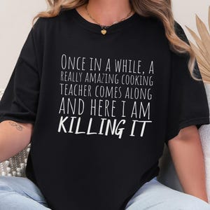 May include: Black t-shirt with white text that reads: "ONCE IN A WHILE, A REALLY AMAZING COOKING TEACHER COMES ALONG AND HERE I AM KILLING IT." The shirt has a crew neck and short sleeves.