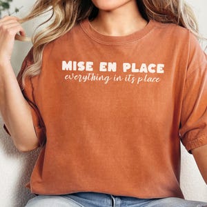 May include: Burnt orange t-shirt with the words "MISE EN PLACE everything in its place" printed in white. The shirt has a crew neck and short sleeves. The fabric appears soft and comfortable.