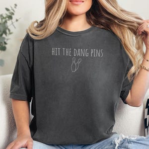 May include: A charcoal gray short-sleeve t-shirt featuring the white text "HIT THE DANG PINS" centered on the chest, accompanied by a simple white line drawing of two bowling pins positioned directly below the printed lettering.
