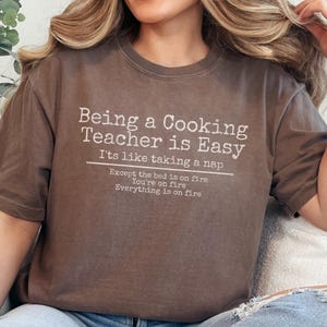 May include: Brown t-shirt with the text "Being a Cooking Teacher is Easy" in white. The shirt has a humorous quote about cooking teachers. The shirt is a casual wear item.