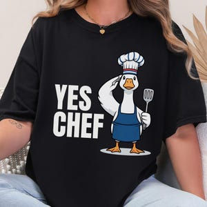 May include: Black t-shirt with the text "YES CHEF" in white. The shirt features a cartoon goose wearing a chef's hat and apron, saluting and holding a spatula. The goose is white with orange feet and a blue apron.