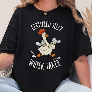 May include: Black t-shirt with a whimsical graphic of a goose wearing a chef's hat and holding whisks. The text reads "CERTIFIED SILLY WHISK TAKER" in white, playful lettering. A fun, food-themed design.
