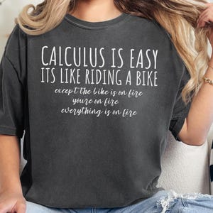 May include: Dark gray t-shirt with the text "CALCULUS IS EASY ITS LIKE RIDING A BIKE except the bike is on fire youre on fire everything is on fire" in white. The shirt is a casual style.