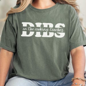 May include: An olive green t-shirt with the text "Dibs on the cooking teacher" in white block letters. The shirt is a short-sleeved crew neck style.