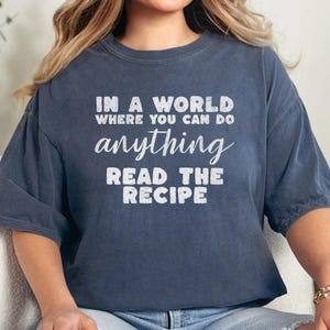 May include: Dark blue t-shirt with the text "IN A WORLD WHERE YOU CAN DO ANYTHING READ THE RECIPE" in white, distressed lettering. The shirt has a relaxed fit and short sleeves. The overall style is casual and comfortable.