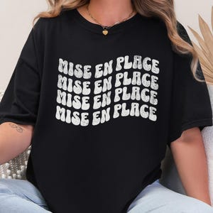 May include: Black t-shirt with the phrase "MISE EN PLACE" in white, wavy, retro-style lettering. The shirt is a crew neck and appears to be made of a soft material.