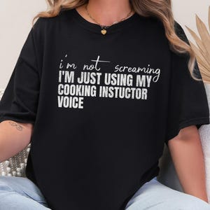 May include: Black t-shirt with white text that reads "I'm not screaming I'm just using my cooking instructor voice". The shirt has a crew neck and short sleeves. The text is in a mix of cursive and block letters.