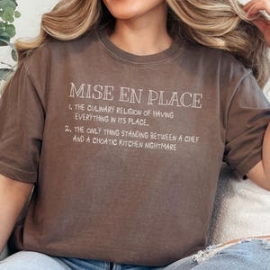 May include: Brown t-shirt with the text "MISE EN PLACE" and two lines of text below. The first line reads "1. THE CULINARY RELIGION OF HAVING EVERYTHING IN ITS PLACE." The second line reads "2. THE ONLY THING STANDING BETWEEN A CHEF AND A CHOATIC KITCHEN NIGHTMARE."