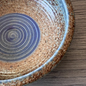 May include: A ceramic bowl featuring a speckled tan and brown textured exterior and rim, transitioning into a smooth, circular purple center with concentric rings etched into the surface, creating a contrast between the rough and polished finish.