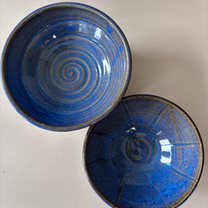 May include: Two round ceramic bowls with a glossy blue glaze and exposed brown clay edges. The top bowl features a spiral pattern etched into the interior, while the bottom bowl displays a spiral center with radiating lines extending to the rim.