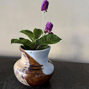 May include: A small, handcrafted ceramic vase with a light blue and brown glaze. The vase holds green leaves and two vibrant purple globe amaranth flowers. The vase is sitting on a dark surface.