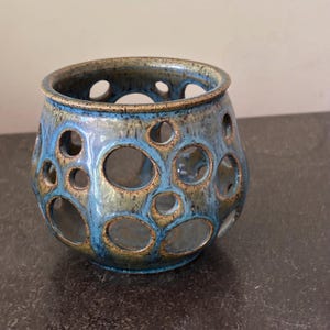 May include: A ceramic teal and blue votive candle holder with a textured finish. The vessel features numerous circular cutouts, creating a decorative and functional design. The rim and edges have a contrasting brown glaze.