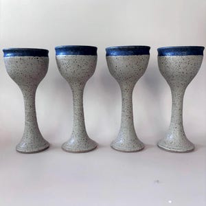 May include: A set of four ceramic goblets, each featuring a speckled light gray body and a smooth, deep blue glazed rim. The goblets have a tall, slender stem and a wide, flared base, maintaining a consistent shape and speckled texture throughout.