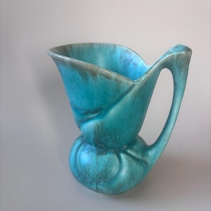 May include: A turquoise ceramic pitcher featuring a bulbous base and a flared, wide rim. The surface displays a mottled, blended finish with darker brown-toned accents throughout the glaze, complemented by a single integrated handle on the side.