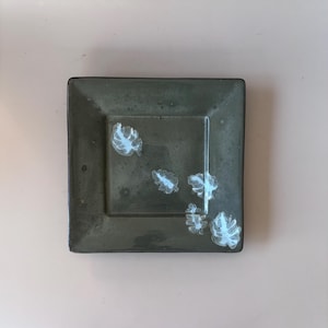 May include: A square ceramic dish with a dark olive green glaze and a smooth, glossy finish. The center of the dish features five small, white, leaf-shaped patterns arranged in a scattered, asymmetrical design across the surface.