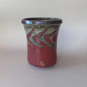 May include: A ceramic tumbler featuring a deep red base glaze and a contrasting upper section with a textured, sage green and dark brown drip pattern. The vessel has a flared rim and a small, circular clay stamp near the base.