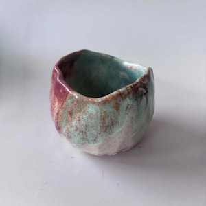 May include: A small, hand-formed ceramic cup with an irregular, organic shape. The exterior and interior feature a mottled glaze in shades of soft turquoise, dusty rose, and earthy brown, creating a blended, textured appearance.