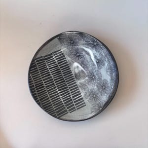 May include: A round ceramic plate featuring a split design. One half displays a pattern of dark gray rectangular dashes on a light gray background, while the other half shows a repeating black and white line drawing of citrus fruit slices.