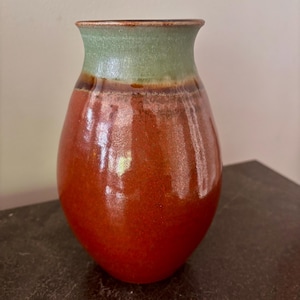 May include: A handmade ceramic vase with a rounded body and a flared rim. The vase features a glossy finish with a gradient of colors, transitioning from a reddish-orange base to a green top. The vase is approximately 8 inches tall.