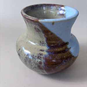 May include: A small ceramic vase with a rounded body and a flared rim. The surface features a glaze with a blend of light blue, cream, and dark brown tones, showing a visible crackle texture throughout the finish of the pottery piece.