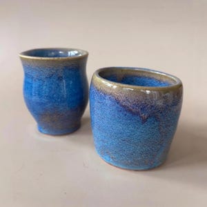 May include: Two ceramic cups with a vibrant blue glaze featuring mottled textures and subtle brown undertones along the rims and bases. The cup on the left has a slightly tapered waist, while the cup on the right has a rounded, cylindrical shape.