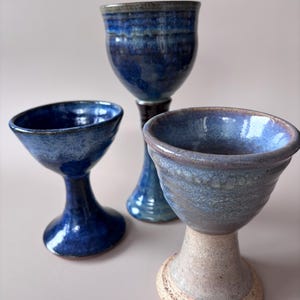 May include: Three ceramic goblets with distinct glazes. One is deep blue with a glossy finish, another is a taller dark blue vessel, and the third features a light speckled beige base transitioning into a mottled blue and purple upper bowl.