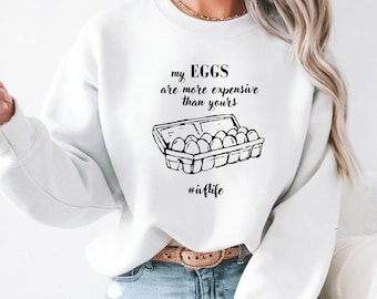 以下が含まれることがあります： 白いスウェットシャツ。黒い卵のカートンのグラフィックと「my EGGS are more expensive than yours」と「#iiflife」のテキスト。カジュアルなスタイル。