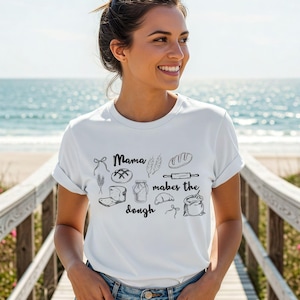 May include: White t-shirt with a black line drawing of bread-making tools and the text "Mama makes the dough." The design includes a rolling pin, bread loaf, and a bag of flour. The shirt is worn by a person on a wooden walkway.