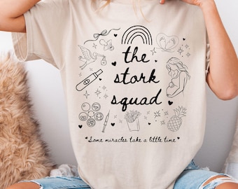 May include: Beige t-shirt with a black graphic design featuring a rainbow, stork, pregnant woman, and the text "the stork squad." The design includes a pacifier, baby feet, and the phrase "Some miracles take a little time."
