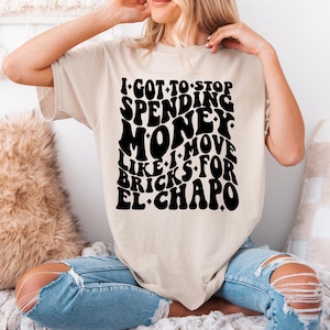 以下が含まれることがあります： 黒いグラフィックプリントが施されたベージュのTシャツ。プリントには「I got to stop spending money like I move bricks for El Chapo」と書いてあります。