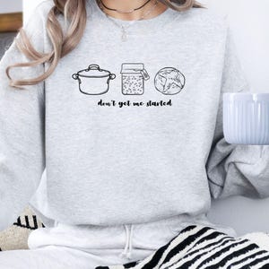 May include: A pale grey sweatshirt featuring a black line drawing of a cooking pot, a jar, and a loaf of bread. The words "don't get me started" are printed below. The sweatshirt is a casual style.