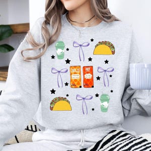 May include: Gray sweatshirt with a print of various Taco Bell items, including tacos, sauce packets, and drinks. The text on the sauce packets reads "(Mild) You're My Fav" and "Hot On The Hot One".