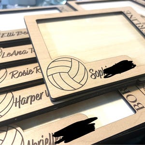 May include: A stack of rectangular light-colored wooden picture frames featuring a laser-engraved volleyball graphic in the corner. Each frame is personalized with a name in script font and the text BORDERLINE Volleyball engraved on the side.