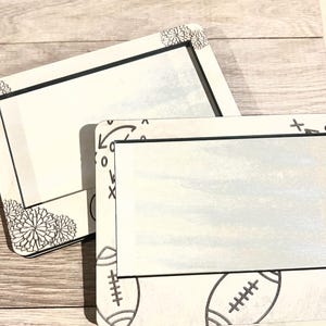 May include: Two white rectangular frames with black trim. One frame has a floral design in the corner, the other has footballs and other graphics. The frames are on a light wood surface.
