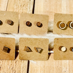 Peut inclure: Présentation de six paires de boucles d'oreilles clous en bois, chacune sur un support en carton brun. Les boucles d'oreilles présentent diverses formes géométriques, notamment des cercles, des carrés et des triangles, dans des tons de bois clairs et foncés. Elles sont présentées sur une surface blanche semblable à de la fourrure.