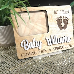May include: Wooden baby announcement frame with the words "Two Little Feet" and baby footprints. The frame includes the name "Baby Williams", "COMING SOON", and "SPRING 2026". The frame is made of light-colored wood.