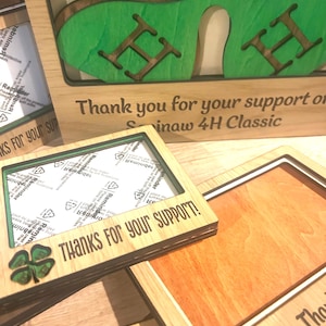 Puede incluir: Una colección de marcos de madera cortados con láser con un diseño de trébol de cuatro hojas verde y el texto grabado "THANKS FOR YOUR SUPPORT!" y "Thank you for your support of the Saginaw 4H Classic." Algunos marcos incluyen película protectora.