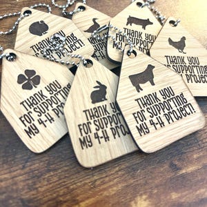 Puede incluir: Una colección de llaveros de madera con forma de casa y cadenas de bolas plateadas. Cada etiqueta presenta un icono de animal o trébol grabado con láser y el texto THANK YOU FOR SUPPORTING MY 4-H PROJECT! La veta de la madera es visible.